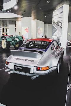 A classic silver Porsche vehicle is displayed in what appears to be a modern car museum. The car is parked on a black, glossy floor, and its rear end is prominently visible, showcasing the Porsche logo and detailing. In the background, a vintage green tractor adds contrast and historical context. The walls feature bright graphic panels and black-and-white photographs, adding visual diversity.