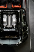 Aerial view of a luxury sports car with a sleek black exterior and red interior accents. The engine layout is prominently displayed, featuring two visible engine components marked with 'EB' and '16.4'. The car's glossy surface reflects lighting, emphasizing its polished finish.
