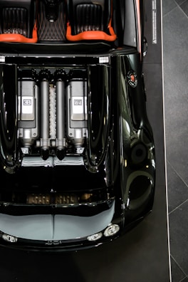 Aerial view of a luxury sports car with a sleek black exterior and red interior accents. The engine layout is prominently displayed, featuring two visible engine components marked with 'EB' and '16.4'. The car's glossy surface reflects lighting, emphasizing its polished finish.