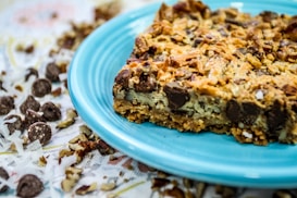 A close-up of a dessert bar featuring chocolate chips, nuts, and coconut flakes, served on a blue plate. The background shows scattered chocolate chips, nuts, and coconut flakes.