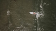 A person paddles on a surfboard in the ocean, with waves creating patterns around them. The view is aerial, capturing the movement of the water and the solitary figure in a dynamic scene.