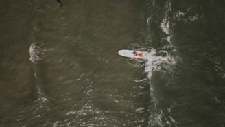 A person paddles on a surfboard in the ocean, with waves creating patterns around them. The view is aerial, capturing the movement of the water and the solitary figure in a dynamic scene.