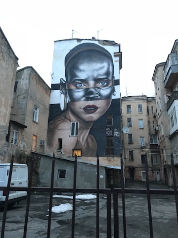 A large mural painted on the side of a building depicting a stylized portrait of a person with metallic tones, striking blue eyes, and bold red lips. The background includes other buildings with a mix of windows and architectural details, creating an urban environment.