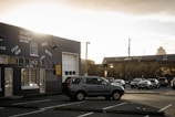 A parking lot is partially filled with cars, including a prominent SUV in the foreground. The scene is set in an urban area, with a large industrial-looking building on the left showcasing signage for an estate liquidators business offering cash for goods like bikes, movies, vinyl, gold, and silver. In the background, a larger building with a flat roof is visible under a cloudy, overcast sky that is illuminated by hues of a setting or rising sun.