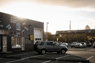 A parking lot is partially filled with cars, including a prominent SUV in the foreground. The scene is set in an urban area, with a large industrial-looking building on the left showcasing signage for an estate liquidators business offering cash for goods like bikes, movies, vinyl, gold, and silver. In the background, a larger building with a flat roof is visible under a cloudy, overcast sky that is illuminated by hues of a setting or rising sun.