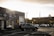 A parking lot is partially filled with cars, including a prominent SUV in the foreground. The scene is set in an urban area, with a large industrial-looking building on the left showcasing signage for an estate liquidators business offering cash for goods like bikes, movies, vinyl, gold, and silver. In the background, a larger building with a flat roof is visible under a cloudy, overcast sky that is illuminated by hues of a setting or rising sun.