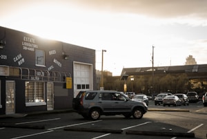 A parking lot is partially filled with cars, including a prominent SUV in the foreground. The scene is set in an urban area, with a large industrial-looking building on the left showcasing signage for an estate liquidators business offering cash for goods like bikes, movies, vinyl, gold, and silver. In the background, a larger building with a flat roof is visible under a cloudy, overcast sky that is illuminated by hues of a setting or rising sun.