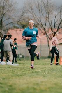 A woman is jogging outdoors on a grassy field surrounded by a group of people. She appears to be smiling and wearing a blue athletic shirt and black leggings. Some cones and what seems to be sports equipment are visible in the background. The sky is overcast, and there are barren trees in the background.