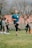 A woman is jogging outdoors on a grassy field surrounded by a group of people. She appears to be smiling and wearing a blue athletic shirt and black leggings. Some cones and what seems to be sports equipment are visible in the background. The sky is overcast, and there are barren trees in the background.