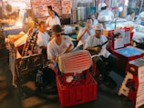 A lively street market scene with a group of musicians playing traditional instruments. The musicians, dressed casually, are seated in a line with various string and percussion instruments. Stalls in the background have colorful fabrics and other items, creating a vibrant atmosphere enhanced by warm lighting. People are browsing the stalls, adding to the bustling ambiance.