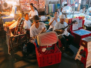 A lively street market scene with a group of musicians playing traditional instruments. The musicians, dressed casually, are seated in a line with various string and percussion instruments. Stalls in the background have colorful fabrics and other items, creating a vibrant atmosphere enhanced by warm lighting. People are browsing the stalls, adding to the bustling ambiance.