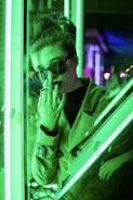 A person wearing sunglasses and a trench coat stands near a glass surface, illuminated by bright green neon lights. The individual's hand is partially covering their face, creating a mysterious and contemplative atmosphere within the urban setting.