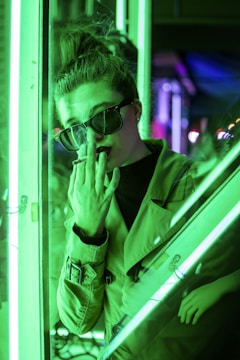 A person wearing sunglasses and a trench coat stands near a glass surface, illuminated by bright green neon lights. The individual's hand is partially covering their face, creating a mysterious and contemplative atmosphere within the urban setting.