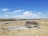 A wooden wagon loaded with logs is positioned on a gravel area in an open grassy landscape. The sky is clear with a few white clouds scattered across it. In the background, a railway track runs parallel to the horizon, with distant hills and sparse vegetation.