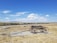 A wooden wagon loaded with logs is positioned on a gravel area in an open grassy landscape. The sky is clear with a few white clouds scattered across it. In the background, a railway track runs parallel to the horizon, with distant hills and sparse vegetation.
