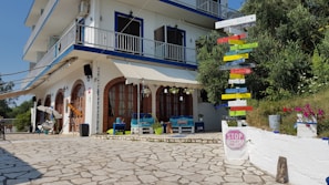 A multi-story building with white walls and blue accents stands on a cobblestone patio. The lower floor features arched windows and various seating arrangements, including colorful cushions and potted plants. A prominent signpost with colorful directional arrows indicates various world cities. The area is complemented by green shrubs and bright flowers, creating a lively and welcoming atmosphere.