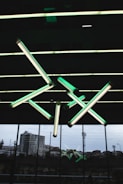 A geometric arrangement of fluorescent light tubes hangs from a ceiling, emitting a greenish glow. The background features large glass windows with an urban landscape visible outside, including buildings and stadium lights.