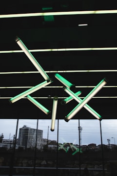 A geometric arrangement of fluorescent light tubes hangs from a ceiling, emitting a greenish glow. The background features large glass windows with an urban landscape visible outside, including buildings and stadium lights.