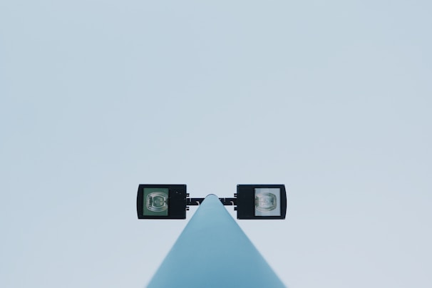A modern light pole with two square floodlights extending to either side against a clear blue sky. The pole is centrally aligned and viewed from a low angle, emphasizing the symmetry and minimalistic design.