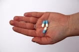 A hand is holding three capsules that are half blue and half white, set against a plain white background. The person is wearing a silver ring on the ring finger.