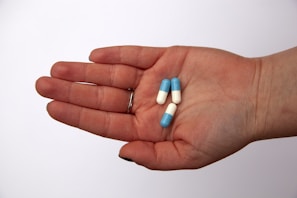 A hand is holding three capsules that are half blue and half white, set against a plain white background. The person is wearing a silver ring on the ring finger.