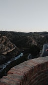 A scenic landscape with a winding road weaving through rugged, rocky hills. The road is bordered by a low brick wall in the foreground. A river or stream is seen cutting through the valley, and a few cars are visible on the road. The sky is clear, adding to the serene and tranquil atmosphere of the scene.