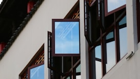 Open windows are part of a modern building design that features clear glass panels and dark-colored frames. The reflection of the sky is visible in the glass, adding a vibrant touch to the structure. The architecture includes clean lines and a minimalist aesthetic with some traditional influences noticeable in the window design pattern.