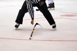 A referee on an ice hockey rink bends over to pick up a broken hockey stick. The player wears black pants, a black and white striped jersey, and skates. The ice surface is marked with red lines and circles.