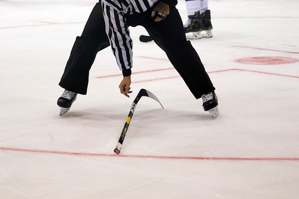 A referee on an ice hockey rink bends over to pick up a broken hockey stick. The player wears black pants, a black and white striped jersey, and skates. The ice surface is marked with red lines and circles.