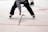 A referee on an ice hockey rink bends over to pick up a broken hockey stick. The player wears black pants, a black and white striped jersey, and skates. The ice surface is marked with red lines and circles.