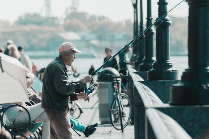 An elderly man is engaged in fishing, holding a fishing rod while standing near a railing by the water. Several people are seated on benches nearby, and a bicycle is parked close by. The scene is set in an outdoor location, likely a waterfront promenade.