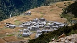 A small village composed of clustered stone buildings sits surrounded by expansive fields. The landscape is rural and serene, with a backdrop of dense forest on the hillsides. Agricultural plots are interspersed among the fields, showing a patchwork of earthy colors.