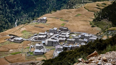 A small village composed of clustered stone buildings sits surrounded by expansive fields. The landscape is rural and serene, with a backdrop of dense forest on the hillsides. Agricultural plots are interspersed among the fields, showing a patchwork of earthy colors.