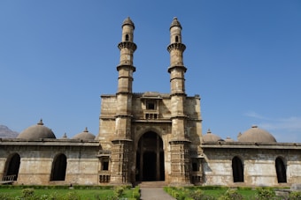 An ancient stone structure with two tall, intricately designed towers flanking a central archway. The building extends sideways with multiple domes visible along the roofline. The architecture includes detailed carvings and embellishments, with a well-maintained lawn in the foreground. The sky is clear and blue, adding a serene backdrop.