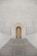 A grand staircase leads up to a large, wooden arched door set into a textured stone wall. The staircase is flanked by rows of vertical, evenly spaced stone columns that create a sense of symmetry and grandeur. The door is ornate, with paneling and large handles.
