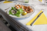 A dining table is set with a white tablecloth and yellow napkins. On a plate, there is a seafood dish that includes shrimp, an oyster on a bed of lettuce, and a lemon wedge. Silverware is neatly placed beside the plate, and wine glasses are visible in the background.