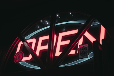 A glowing neon sign displays the word 'OPEN' in bold red letters. The sign is set against a dark background, with a semi-circular shape surrounding it. Small stickers are visible, adding detail to the scene.