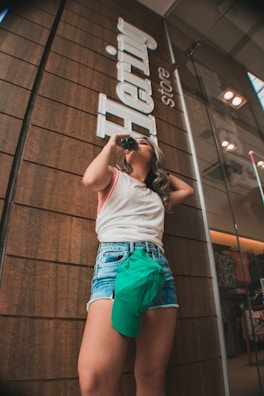 A person stands in front of a store with a wooden panel exterior, sipping from a bottle while wearing casual summer attire. Denim shorts and a sleeveless top are paired with a green cap tucked into the shorts. The store sign reads 'Hering store'.