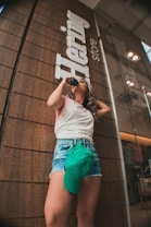 A person stands in front of a store with a wooden panel exterior, sipping from a bottle while wearing casual summer attire. Denim shorts and a sleeveless top are paired with a green cap tucked into the shorts. The store sign reads 'Hering store'.