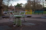 An outdoor playground area with a seesaw featuring green springs and metal handles. The ground is made of soft material, and there's a green park bench and a yellow gate in the background. Surrounding the area are bare trees and some bushes, under a cloudy sky.