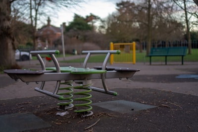 An outdoor playground area with a seesaw featuring green springs and metal handles. The ground is made of soft material, and there's a green park bench and a yellow gate in the background. Surrounding the area are bare trees and some bushes, under a cloudy sky.