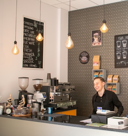 A barista stands behind the counter in a modern coffee shop, smiling. The counter displays coffee cups, a coffee machine, and various equipment and items used in coffee preparation. Overhead, there are three hanging light bulbs with warm lighting. On the wall behind the barista, there are shelves filled with coffee bags, a coffee menu, and other decorative elements.
