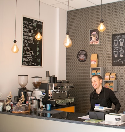 A barista stands behind the counter in a modern coffee shop, smiling. The counter displays coffee cups, a coffee machine, and various equipment and items used in coffee preparation. Overhead, there are three hanging light bulbs with warm lighting. On the wall behind the barista, there are shelves filled with coffee bags, a coffee menu, and other decorative elements.