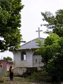 A small white building with a cross on top, likely a church, is nestled among lush green trees. Two children are playing on a rocky path leading to the entrance of the building. The setting appears rural and serene, with natural greenery surrounding the area.