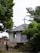 A small white building with a cross on top, likely a church, is nestled among lush green trees. Two children are playing on a rocky path leading to the entrance of the building. The setting appears rural and serene, with natural greenery surrounding the area.