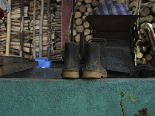 A pair of worn, muddy boots are placed on a teal-colored ledge in front of a background filled with neatly stacked logs. Some farming equipment and a blue container are visible nearby. The setting appears rustic and outdoors, potentially a farm or rural area.