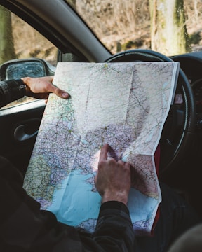 A person holds a detailed map while sitting in a car. The map is open, and one finger is pointing to a specific location. The background outside the car window shows trees and a blurred landscape.