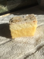 A block of cheese with a rough, crumbly edge and a creamy yellow interior rests on a textured white towel, illuminated by natural light.