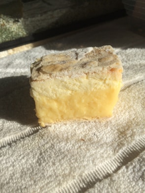 A block of cheese with a rough, crumbly edge and a creamy yellow interior rests on a textured white towel, illuminated by natural light.