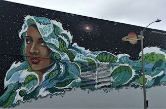 A vibrant mural featuring a woman's face with long, flowing hair that transforms into ocean waves, dotted with phrases like 'beauty of the wild' and 'be with me.' The backdrop is a starry night sky with celestial bodies, including a galaxy and a planet. A streetlamp stands in the foreground.